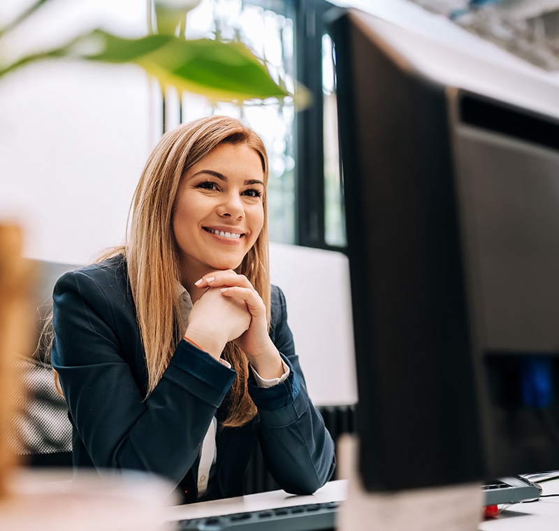 A medical front office administration specialist welcomes a patient at the reception desk, entering information into a computer and managing appointment scheduling in a healthcare office.